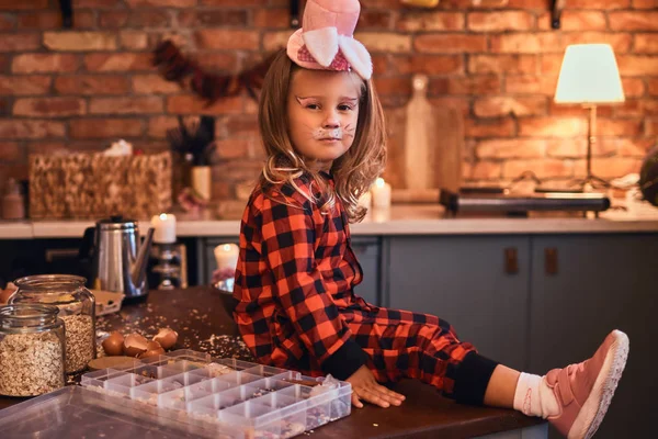 Menina bonito em chapéu de maquiagem de coelho e pijama sentado na mesa com comida espalhada na cozinha estilo loft de manhã . — Fotografia de Stock