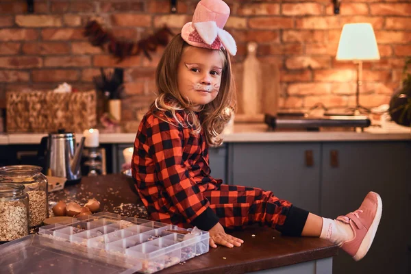 Милая маленькая девочка в кроличьей шляпе и пижаме, сидящая на столе с разбросанной едой в лофт стиле кухни по утрам . — стоковое фото