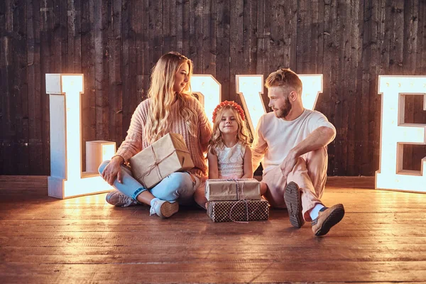 Mamma, Pappa och dotter sitter tillsammans med gåvor i rum inredda med voluminösa bokstäver med belysning. — Stockfoto
