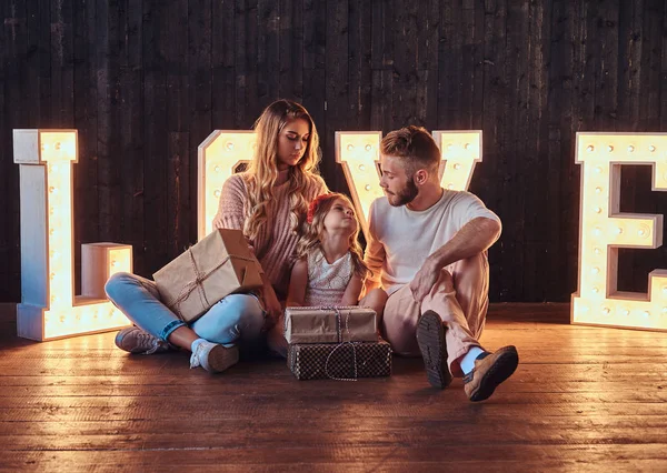 Mamá, papá e hija se sientan juntos con regalos en la habitación decorada con letras voluminosas con iluminación . — Foto de Stock