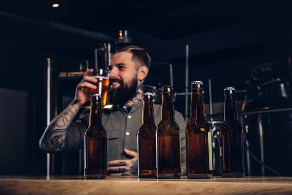 Foto s craft pivních lahví na popředí a vousatý muž pití na pozadí. — Stock fotografie