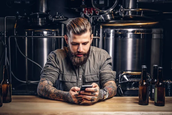Татуйований чоловік-хіпстер зі стильною бородою та волоссям, використовуючи смартфон, сидячи за барною стійкою в пивоварні Інді . — стокове фото