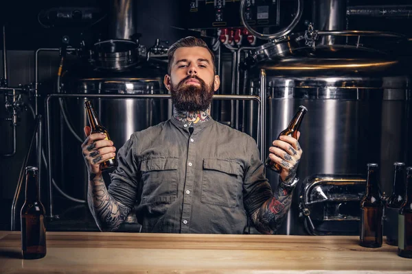 Retrato de un hombre hipster tatuado pensativo con barba y cabello elegantes sostienen dos botellas con cerveza artesanal en la fábrica de cerveza indie . —  Fotos de Stock