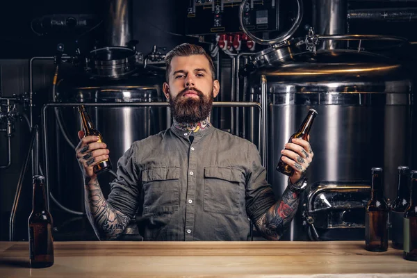 Retrato de um homem hipster tatuado pensivo com barba e cabelo elegantes segurar duas garrafas com cerveja artesanal na cervejaria indie . — Fotografia de Stock