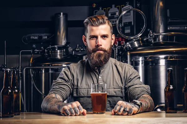 Portret van een man van de getatoeëerde hipster met stijlvolle baard en haren in shirt zitten aan de bar teller met glas bier in indie brouwerij. — Stockfoto