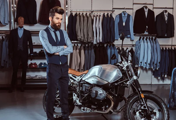 Elegantemente vestito uomo posa con le braccia incrociate vicino moto sportiva retrò presso il negozio di abbigliamento uomo . — Foto Stock