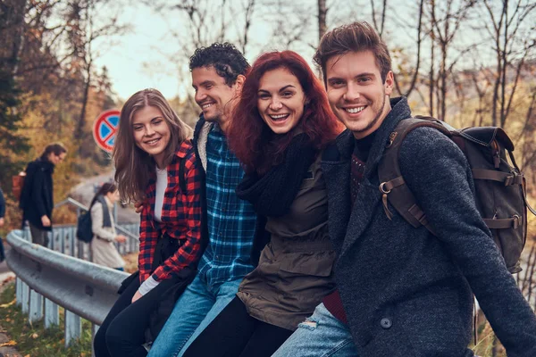 Grupo de jóvenes amigos con mochilas sentados en la barandilla cerca de la carretera con un hermoso bosque y río en el fondo . — Foto de Stock