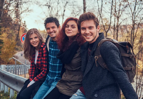 Grupo de jóvenes amigos con mochilas sentados en la barandilla cerca de la carretera con un hermoso bosque y río en el fondo . — Foto de Stock