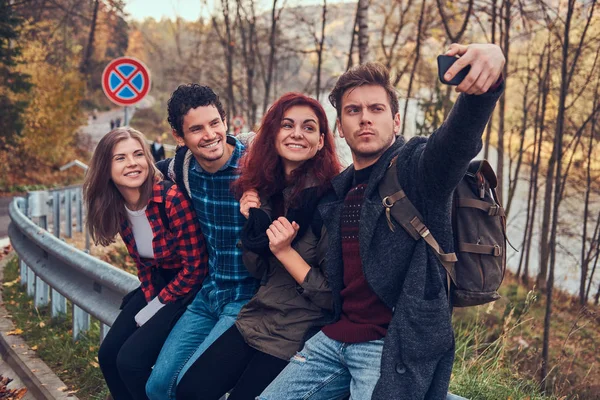Grupo de jóvenes excursionistas con mochilas sentados en la barandilla cerca de la carretera y haciendo selfie . — Foto de Stock