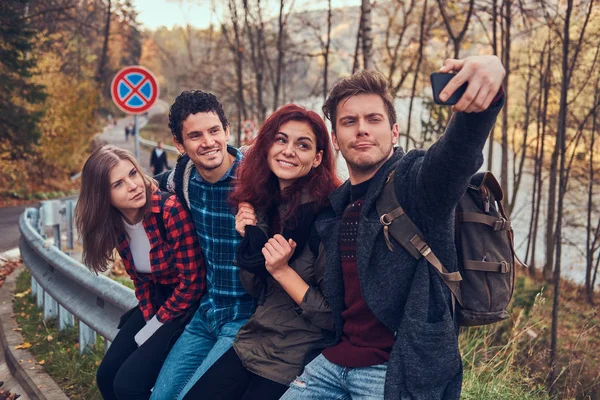 Grupo de jóvenes excursionistas con mochilas sentados en la barandilla cerca de la carretera y haciendo selfie . — Foto de Stock