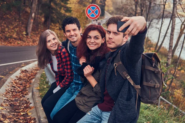 Grupo de jóvenes excursionistas con mochilas sentados en la barandilla cerca de la carretera y haciendo selfie . — Foto de Stock