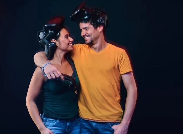 Young couple of gamers wearing virtual reality goggles hug and look at each other. — Stock Photo, Image