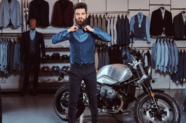 Stylish man dressed in elegant wear posing with hands on bow tie near retro sports motorbike at the mens clothing store. clipart