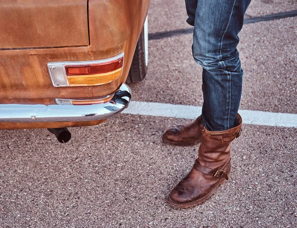 Foto recortada de un hombre en jeans y botas de pie cerca de coche retro en el aparcamiento de la ciudad . — Foto de Stock