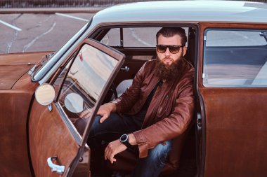 Kahverengi deri ceket giymiş güneş gözlüğü sakallı erkek açık kapı ile ayarlanmış bir retro arabanın direksiyonuna oturur.