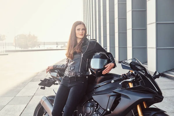 A beautiful biker girl leaning on her superbike outside a building on a sunny day. — Stock Photo, Image