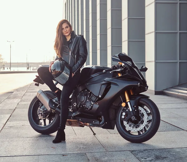 A beautiful biker girl leaning on her superbike outside a building. — Stock Photo, Image