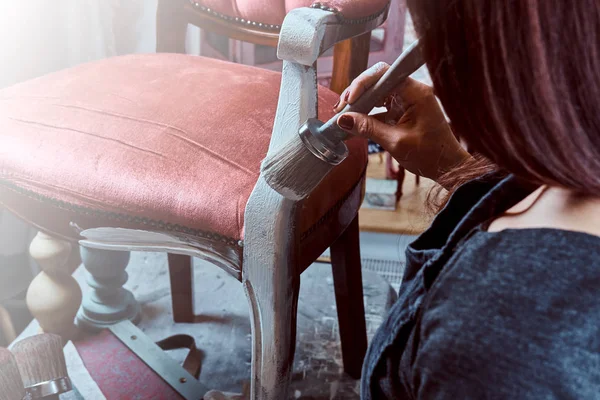 Female artist painting vintage chair in white color with paintbrush in workshop.