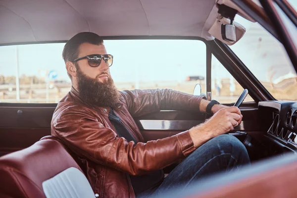 Bebaarde man in zonnebril gekleed in bruine lederen jas retro autorijden. — Stockfoto