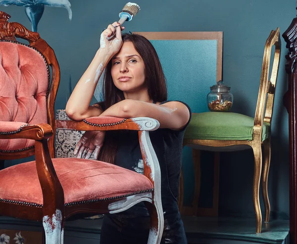 Stanco artista donna in grembiule tiene pennello e appoggiato su una sedia vintage in officina . — Foto Stock