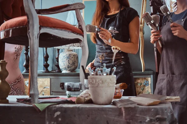 Художница с ассистенткой в фартуках, рисует винтажное кресло кисточкой в мастерской . — стоковое фото