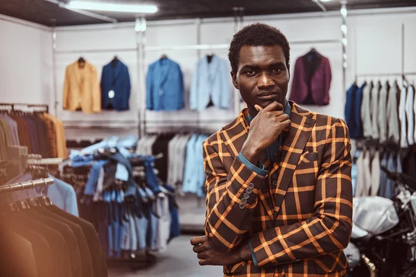 Элегантно одетый африканский мужчина позирует с рукой на подбородке, стоя в классическом магазине мужской одежды . — стоковое фото