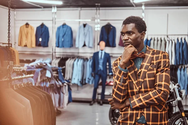Hombre africano elegante vestido posando con la mano en la barbilla mientras está de pie en una tienda de ropa masculina clásica . — Foto de Stock