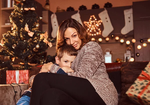 Mom hugging her cute little boy while sitting on sofa in decorated room during Christmas time.