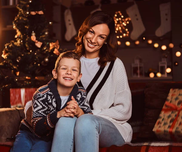 Mama umarmt ihren süßen kleinen Jungen, während sie während der Weihnachtszeit auf dem Sofa im dekorierten Zimmer sitzt. — Stockfoto