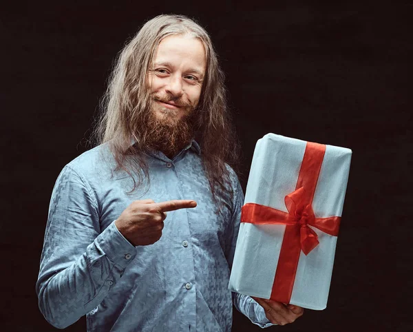 Happy male with long hair and beard in blue shirt pointing on gift box in his hand. Christmas concept.