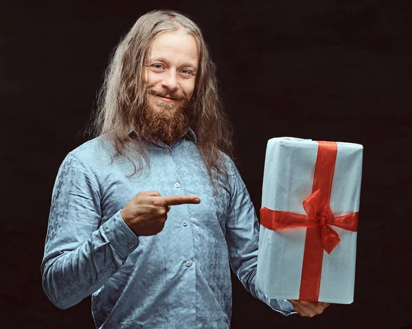 Homem feliz com cabelos longos e barba em camisa azul apontando na caixa de presente em sua mão. Conceito de Natal . — Fotografia de Stock