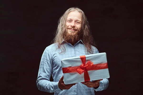 Щасливий чоловік з довгим волоссям і бородою в синій сорочці тримає подарункову коробку. Ізольовані на темному тлі . — стокове фото