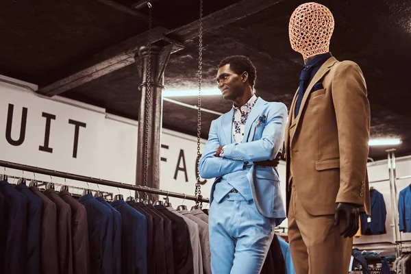 Elegantemente vestito uomo afro-americano in posa con le braccia incrociate vicino manichino in un negozio di abbigliamento maschile classico . — Foto Stock