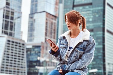 Yüzünde bulutlu sabah Moskova şehir içinde gökdelenler önünde oturan bir smartphone kullanarak denim ceket giyen dövmeli şık Kızıl saçlı hippi kız.