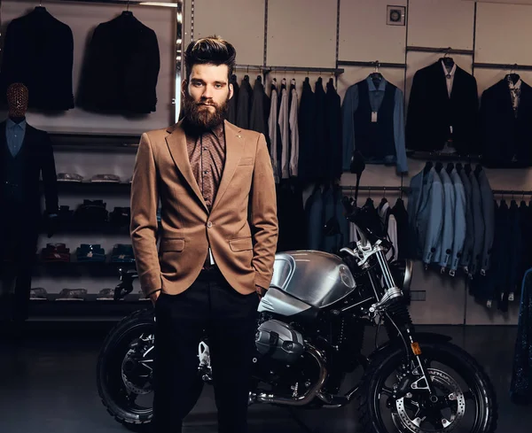 Elegantly dressed man with stylish beard and hair posing near retro sports motorbike at the mens clothing store.