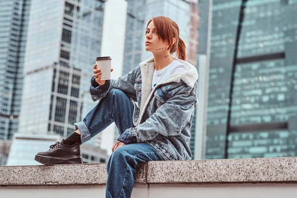 Elegante chica pelirroja hipster con tatuaje en la cara con abrigo de mezclilla sosteniendo café para llevar sentado frente a rascacielos en la ciudad de Moskow en la mañana nublada . —  Fotos de Stock