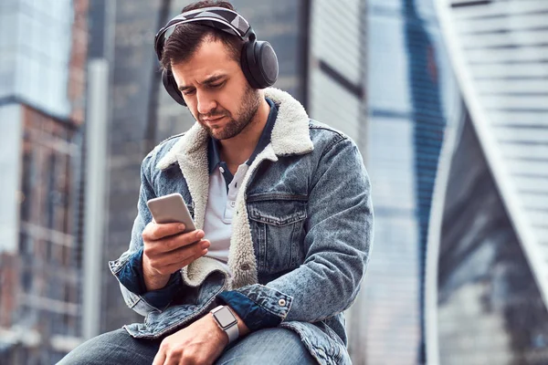 Stilig mann med dongerijakke som lytter til musikk og bruker smarttelefon foran skyskrapere i Moskow City på overskyet morgen . – stockfoto