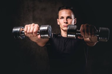 Koyu arka plan üzerinde dumbbells ile egzersiz yaparak spor giyim giyen genç bir vücut geliştirmeci.