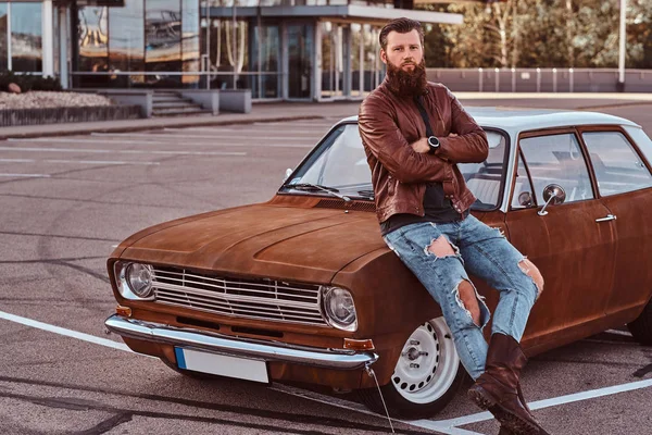 Bärtiger Mann in brauner Lederjacke und Stiefeln lehnt auf getuntem Retro-Auto auf dem Stadtparkplatz in der Nähe von Wolkenkratzer. — Stockfoto