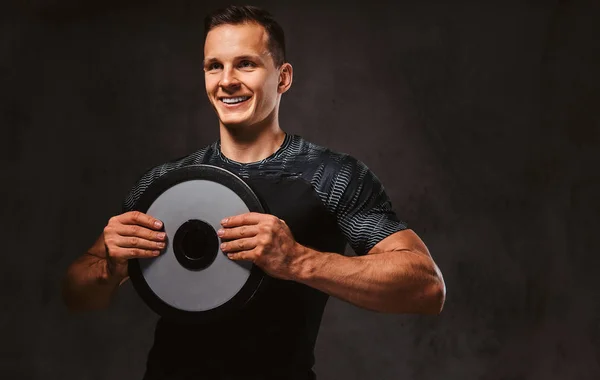 Een jonge man in sportkleding met een barbell schijf op donkere achtergrond. — Stockfoto