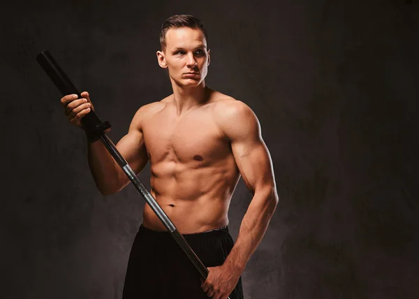 Portrait d'un beau jeune bodybuilder tenant un haltère et regardant loin sur fond sombre . — Photo