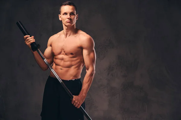 Portrait of a handsome young bodybuilder holding a barbell and looking away on dark background. — Stock Photo, Image