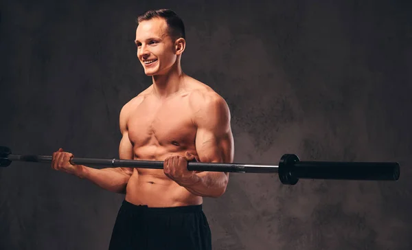 Jonge shirtless bodybuilder holding een barbell en doen oefening op de biceps. Geïsoleerd op donkere achtergrond. — Stockfoto