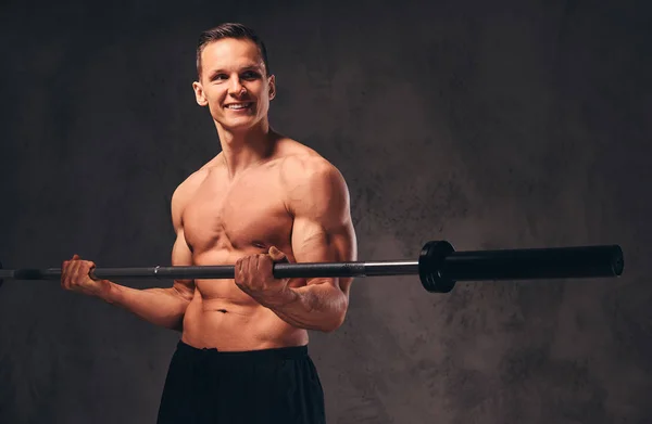Jonge shirtless bodybuilder holding een barbell en doen oefening op de biceps. Geïsoleerd op donkere achtergrond. — Stockfoto