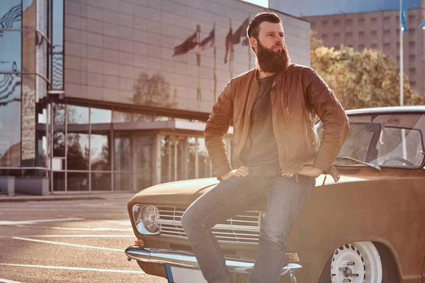 Stylový muž opíraje se o laděný retro auto ve městě, parkování poblíž mrakodrap na světlé západ slunce. — Stock fotografie