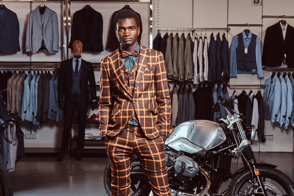 Афро-американец, одетый в модный элегантный костюм, позирует с руками в карманах возле магазина мужской одежды возле ретро-спортивного мотоцикла . — стоковое фото