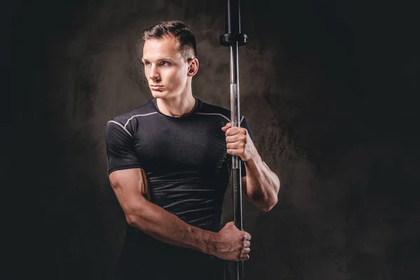 Portret van een knappe jonge bodybuilder houden een barbell en weg op donkere achtergrond. — Stockfoto