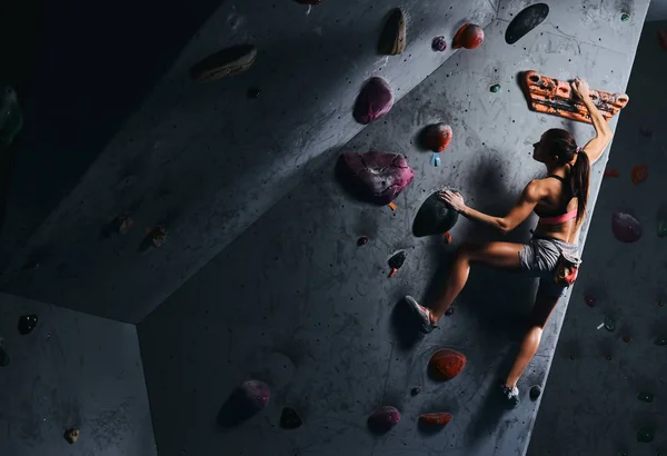 Mujer joven en pantalones cortos y sujetador deportivo haciendo ejercicio en una pared de bouldering en el interior . — Foto de Stock