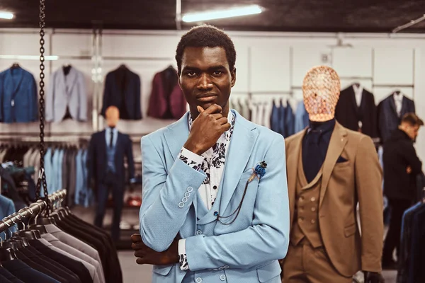 Elegantemente vestito uomo africano in posa con mano sul mento mentre in piedi in un negozio di abbigliamento maschile classico . — Foto Stock