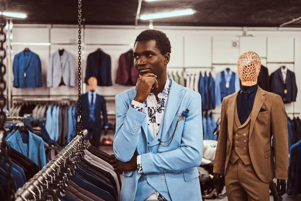 Hombre africano elegante vestido posando con la mano en la barbilla mientras está de pie en una tienda de ropa masculina clásica . — Foto de Stock